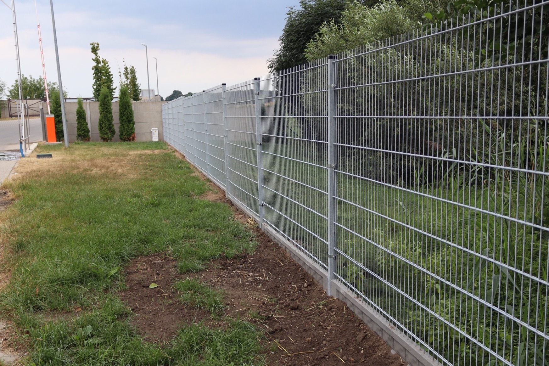 Długi, srebrny ogrodzenie panelowe 2D, zabetonowane u podstawy, rozciąga się wzdłuż trawnika i drzew, z widocznymi słupkami i panelami z drutu.