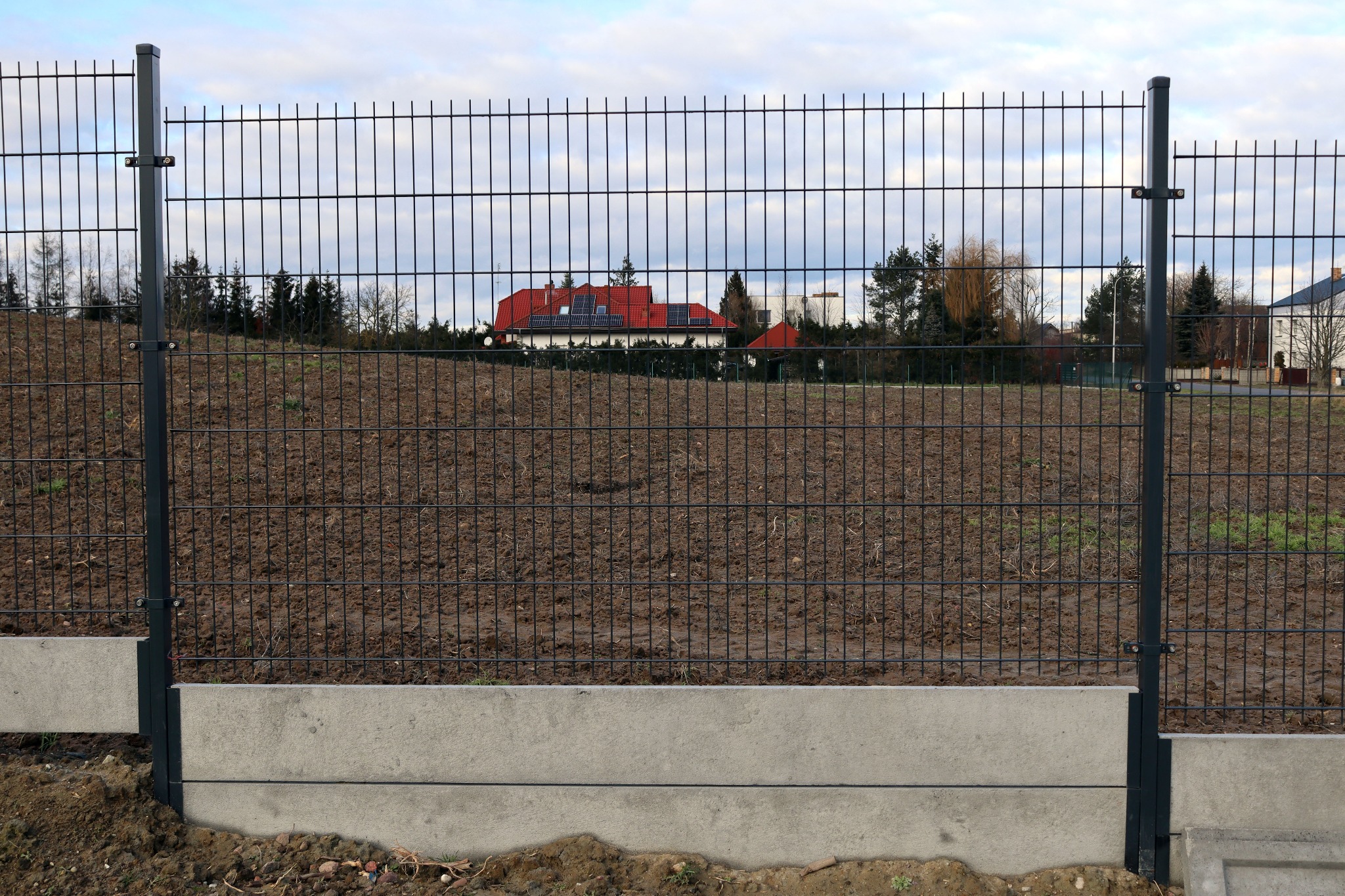 Częściowo zamontowane ogrodzenie panelowe 2D z betonową podmurówką na nierównym terenie, w tle dom z czerwoną dachówką i drzewa.