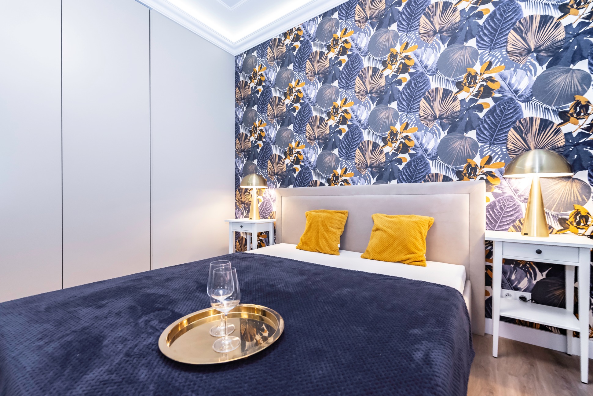 Nowoczesna sypialnia z łóżkiem, złotą tacą z kieliszkami, żółtymi poduszkami i tapetą w tropikalne liście.