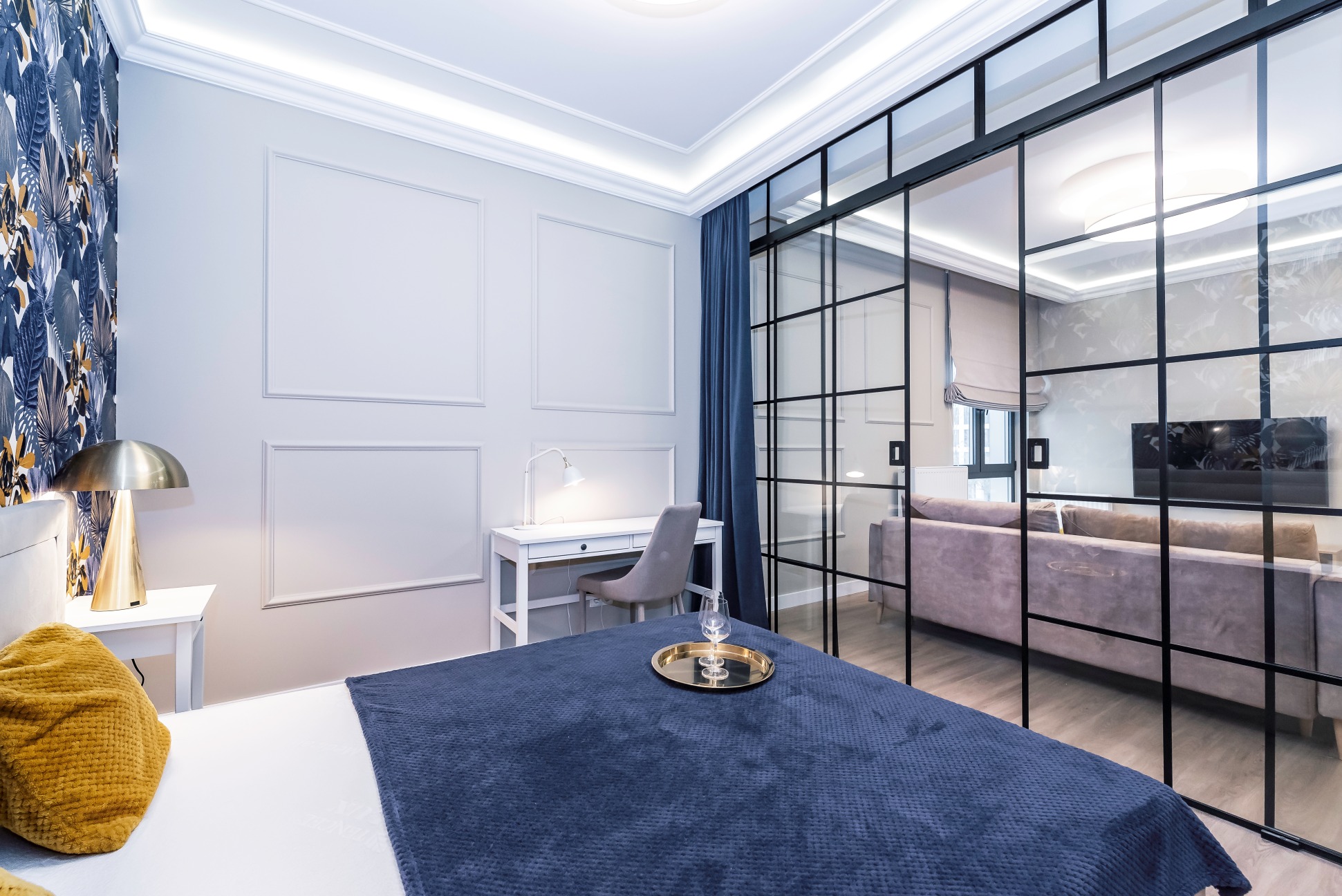 Nowoczesna sypialnia z przeszkloną ścianą działową, biurkiem i tapetą w tropikalne wzory. Na łóżku leży niebieska narzuta i złota taca z kieliszkami.