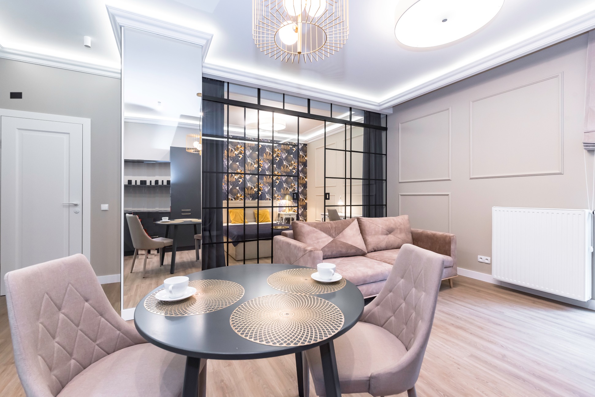 Nowoczesny salon z okrągłym stołem, krzesłami z tapicerką w odcieniach różu, sofą w kolorze pudrowego różu i szklaną ścianką działową z czarnymi ramami oddzielającą sypialnię z tapetą w motywy...