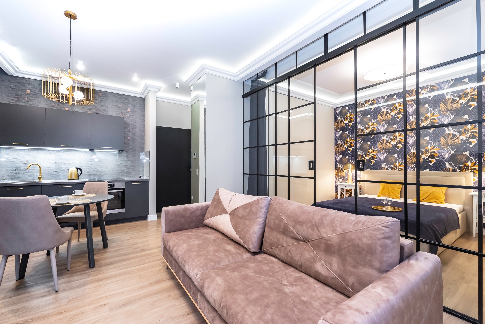 Jasne, nowoczesne wnętrze apartamentu w Warszawie z otwartą kuchnią, salonem i sypialnią oddzieloną szklaną ścianką. Widoczny pluszowy narożnik, okrągły stół z krzesłami oraz dekoracyjna lampa...