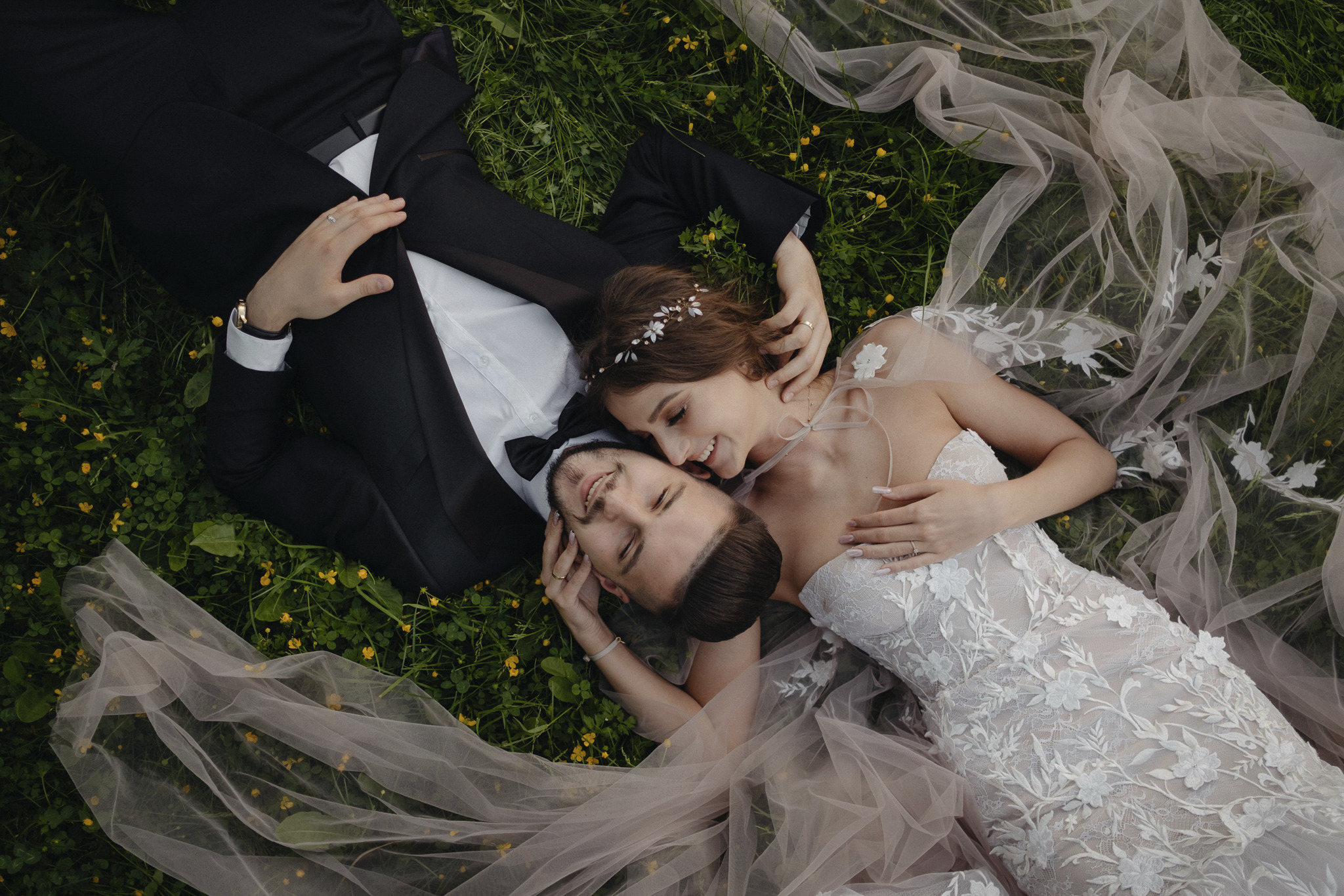 Zakochana para młoda leży na trawie z żółtymi kwiatami, panna młoda w koronkowej sukni z welonem, pan młody w czarnym garniturze z muszką.