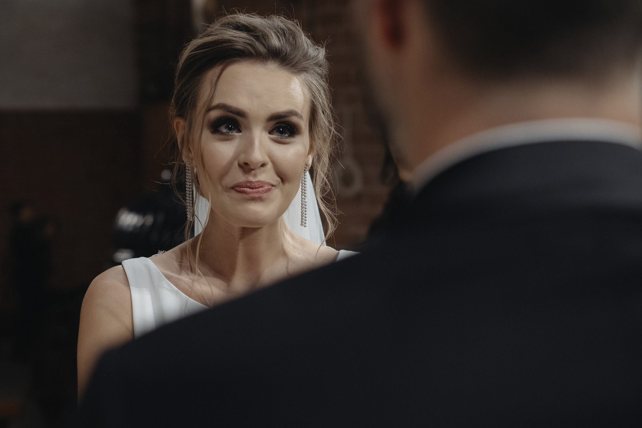 Panna młoda z subtelnym uśmiechem i długimi, błyszczącymi kolczykami, spogląda na pana młodego podczas ceremonii ślubnej.