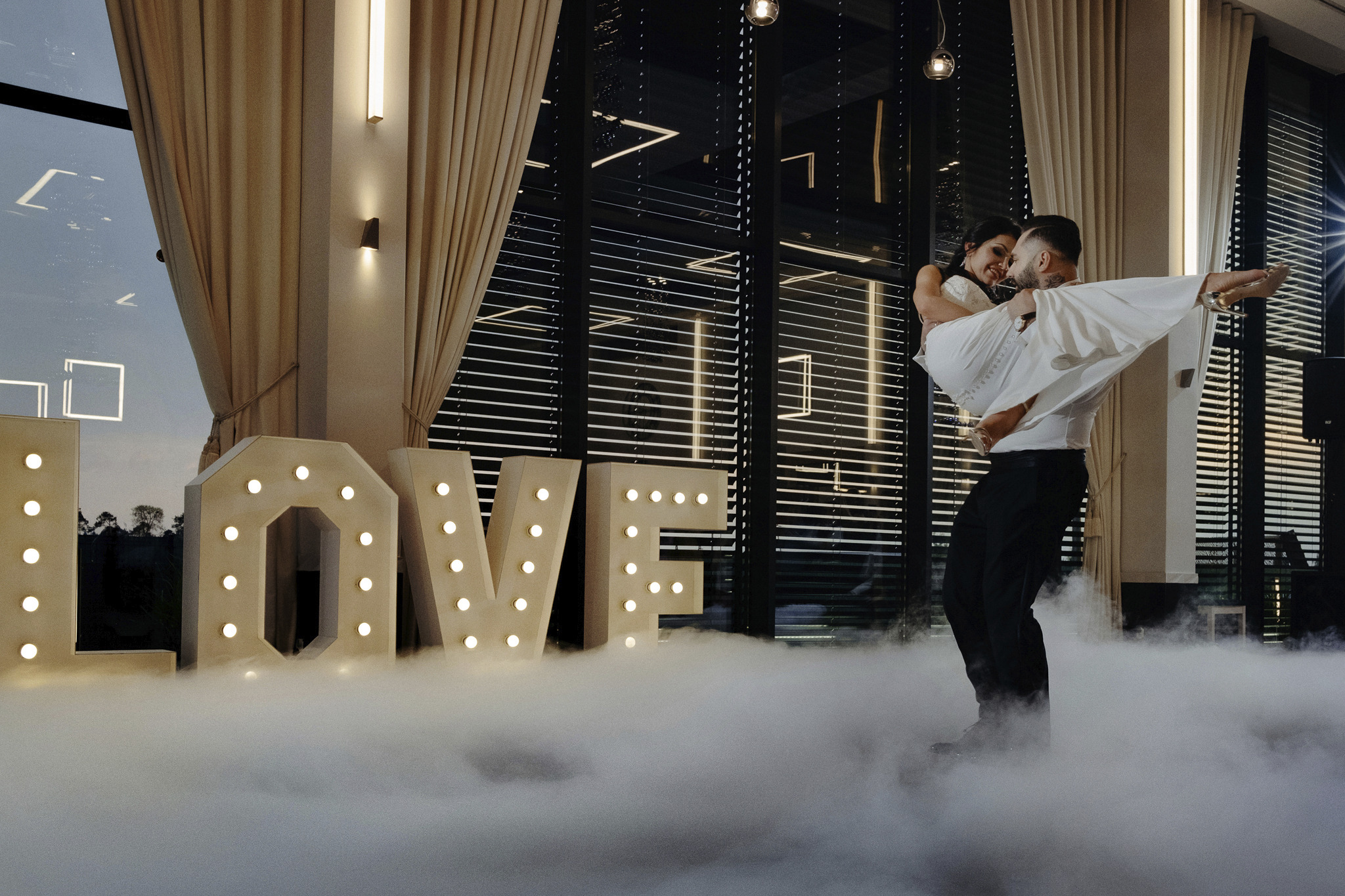 Pan młody trzyma pannę młodą na rękach na tle dużego napisu LOVE z żarówkami, otoczonego sztuczną mgłą, w eleganckim wnętrzu z zasłonami i żaluzjami.