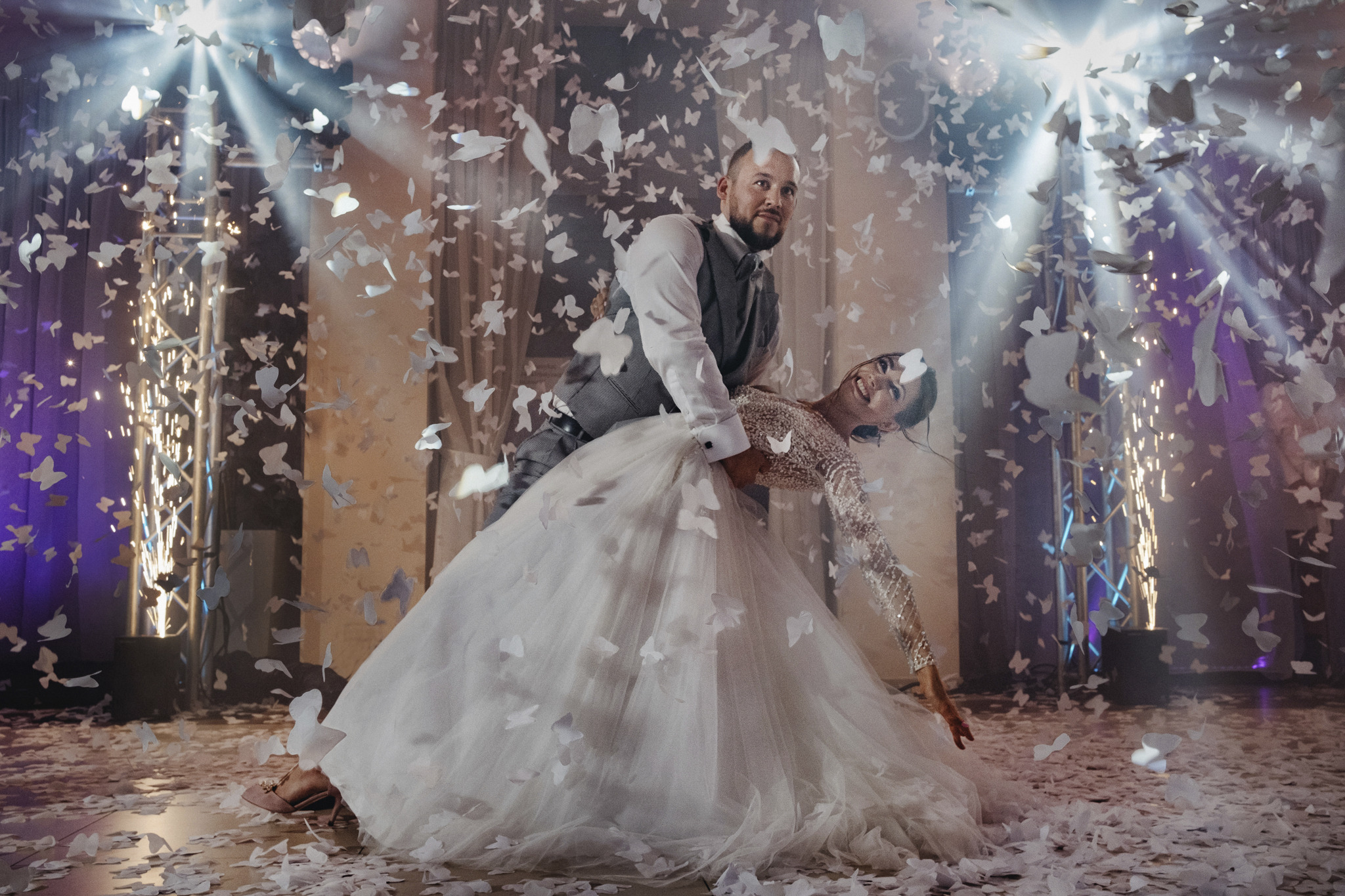 Uśmiechnięta para młoda w tańcu, otoczona konfetti w kształcie motyli i efektami pirotechnicznymi, on w szarej kamizelce, ona w sukni ślubnej z długim rękawem, oświetlenie sceniczne.