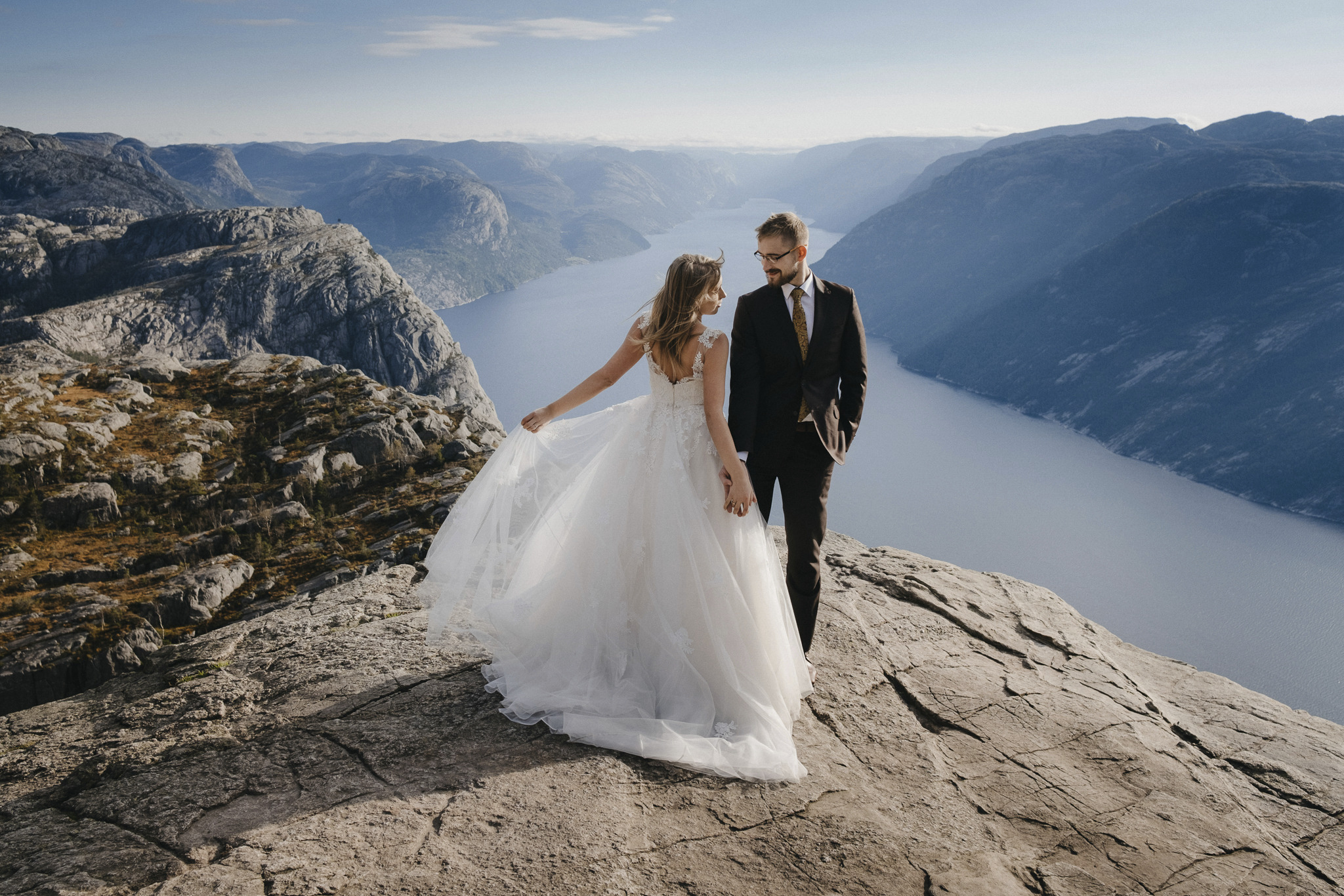 Para młoda na skalistym szczycie z widokiem na fiord, panna młoda w koronkowej sukni z rozkloszowanym dołem, pan młody w ciemnym garniturze i okularach, malowniczy krajobraz w tle.