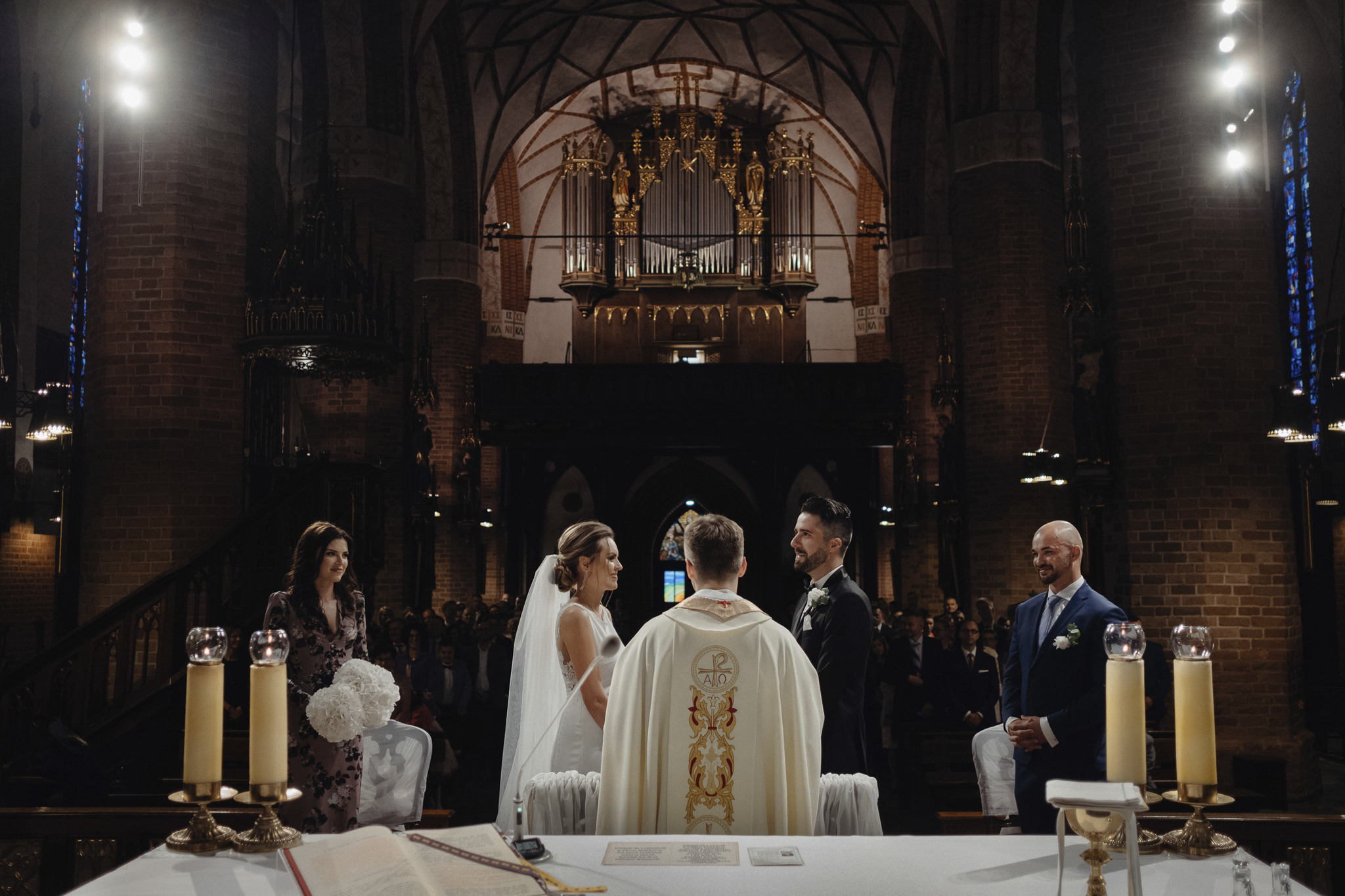 Para młoda podczas ceremonii ślubnej w kościele z organami w tle, w obecności księdza i gości.