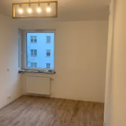 Pusty pokój po remoncie z drewnianą podłogą, białymi ścianami, grzejnikiem pod oknem i nowoczesną lampą sufitową z czterema żarówkami.