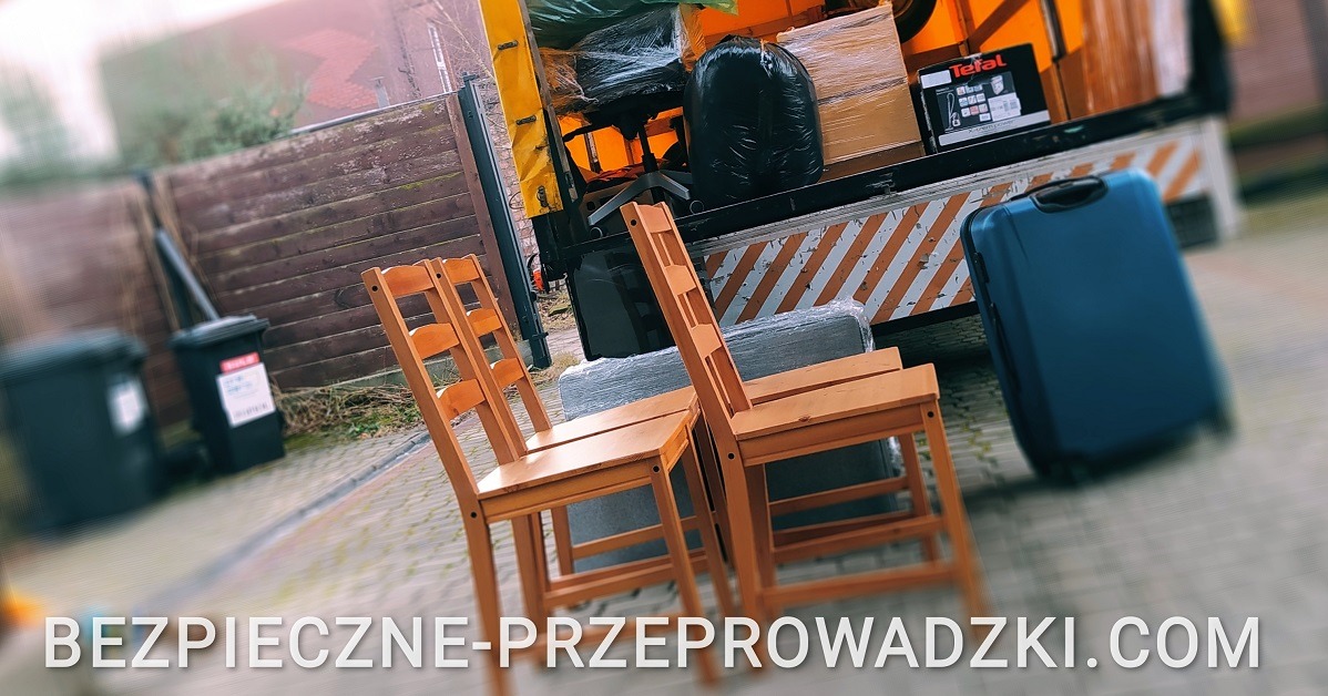 Dwa drewniane krzesła ustawione przed otwartą paką samochodu dostawczego załadowanego meblami i sprzętem AGD, w tle widoczne kosze na śmieci i drewniany płot.