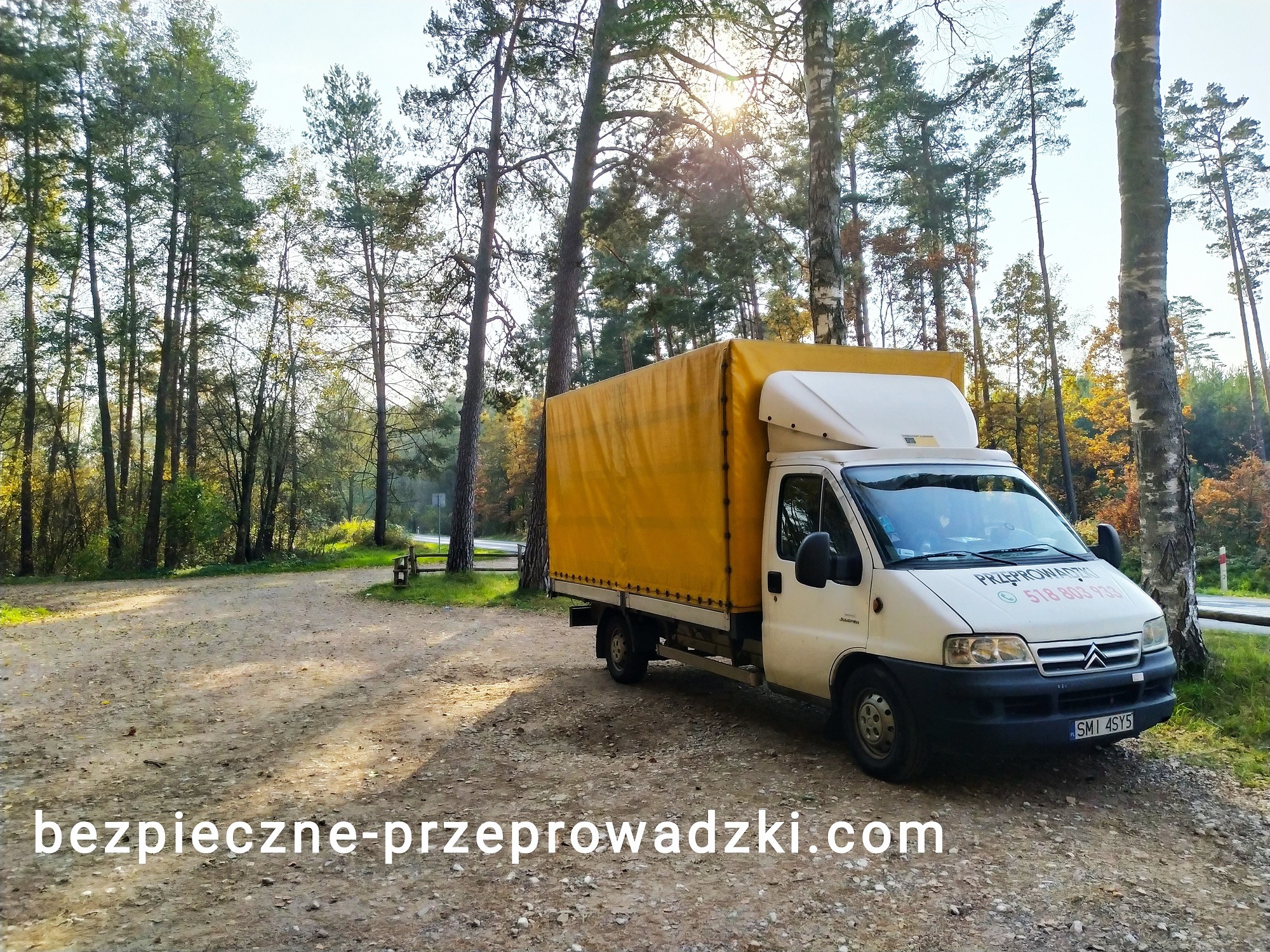 Biały samochód dostawczy z żółtą plandeką zaparkowany na leśnym parkingu, widoczny numer telefonu i napis 'Przeprowadzki' na masce pojazdu.