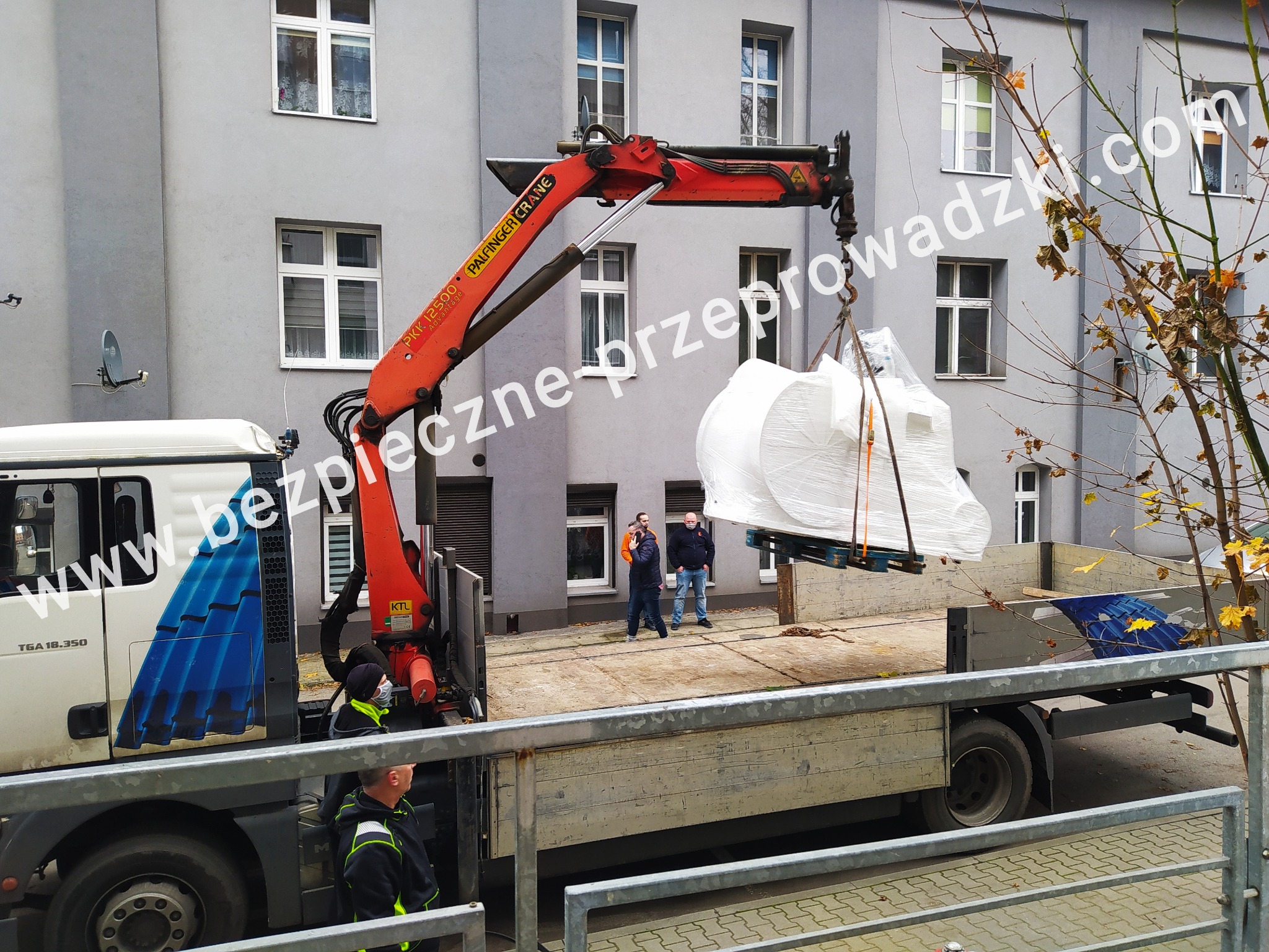 Ciężarówka z dźwigiem Palfinger ładuje owinięty w folię ładunek na palecie, na tle szarego budynku z oknami, widoczni pracownicy w maskach.