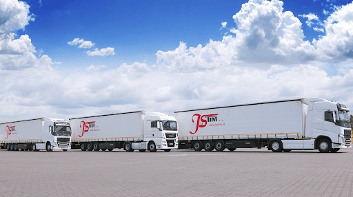 Trzy białe ciężarówki z naczepami ustawione w rzędzie na parkingu, każda z logo firmy JS TIM, na tle błękitnego nieba z chmurami.