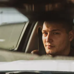 Młody mężczyzna siedzący w samochodzie, widziany przez lusterko wsteczne, z zamyślonym wyrazem twarzy i delikatnym oświetleniem.