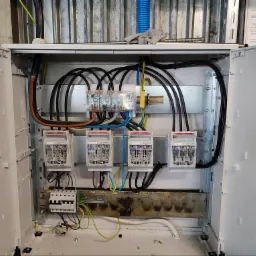 Otwarta skrzynka elektryczna z zainstalowanymi bezpiecznikami, przewodami i listwami zaciskowymi, widoczne ślady przepaleń na listwie dolnej.