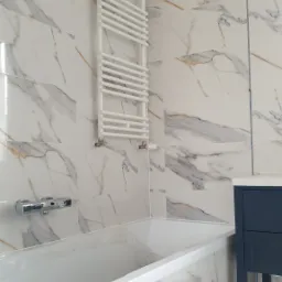 Nowoczesna łazienka z wanną, białą armaturą i grzejnikiem drabinkowym na ścianie wyłożonej płytkami imitującymi marmur.