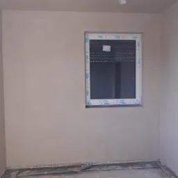 Świeżo nałożone tynki maszynowe na ścianach wewnętrznych pomieszczenia z oknem VEKA z roletami, widoczne ślady po pracach na podłodze.