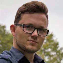 Młody mężczyzna w okularach i granatowej koszuli, z lekkim zarostem, patrzy w obiektyw na tle rozmytej zieleni drzew.