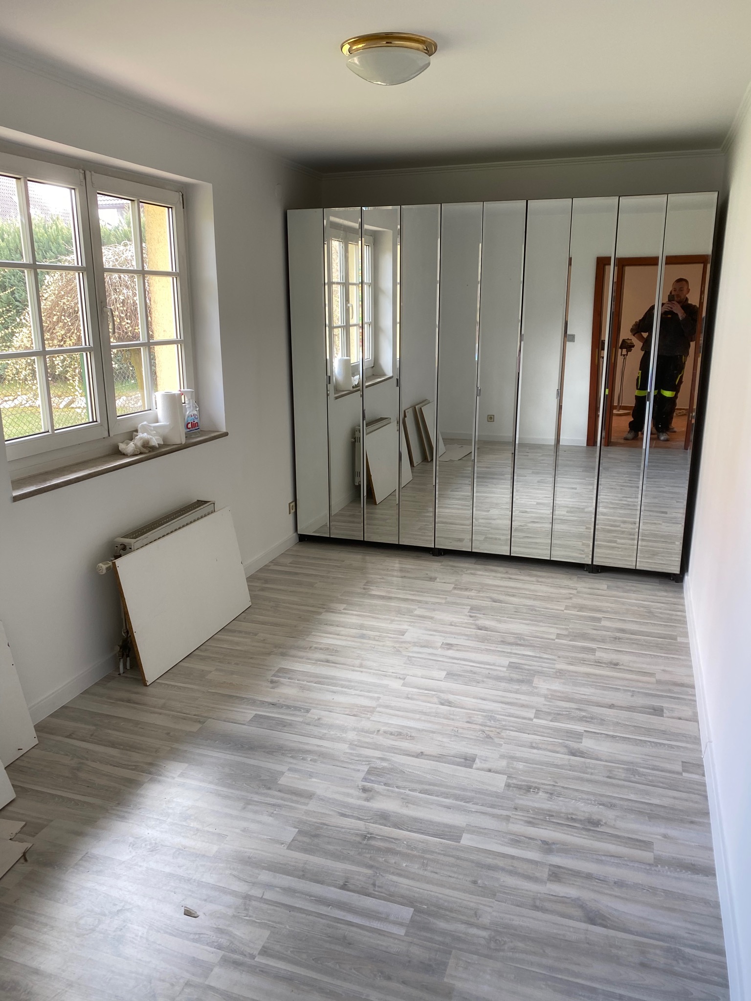 Pokój w trakcie remontu z widoczną ścianą z lustrami, grzejnikiem pod oknem i fragmentami paneli podłogowych. W lustrach odbija się pracownik w kombinezonie.