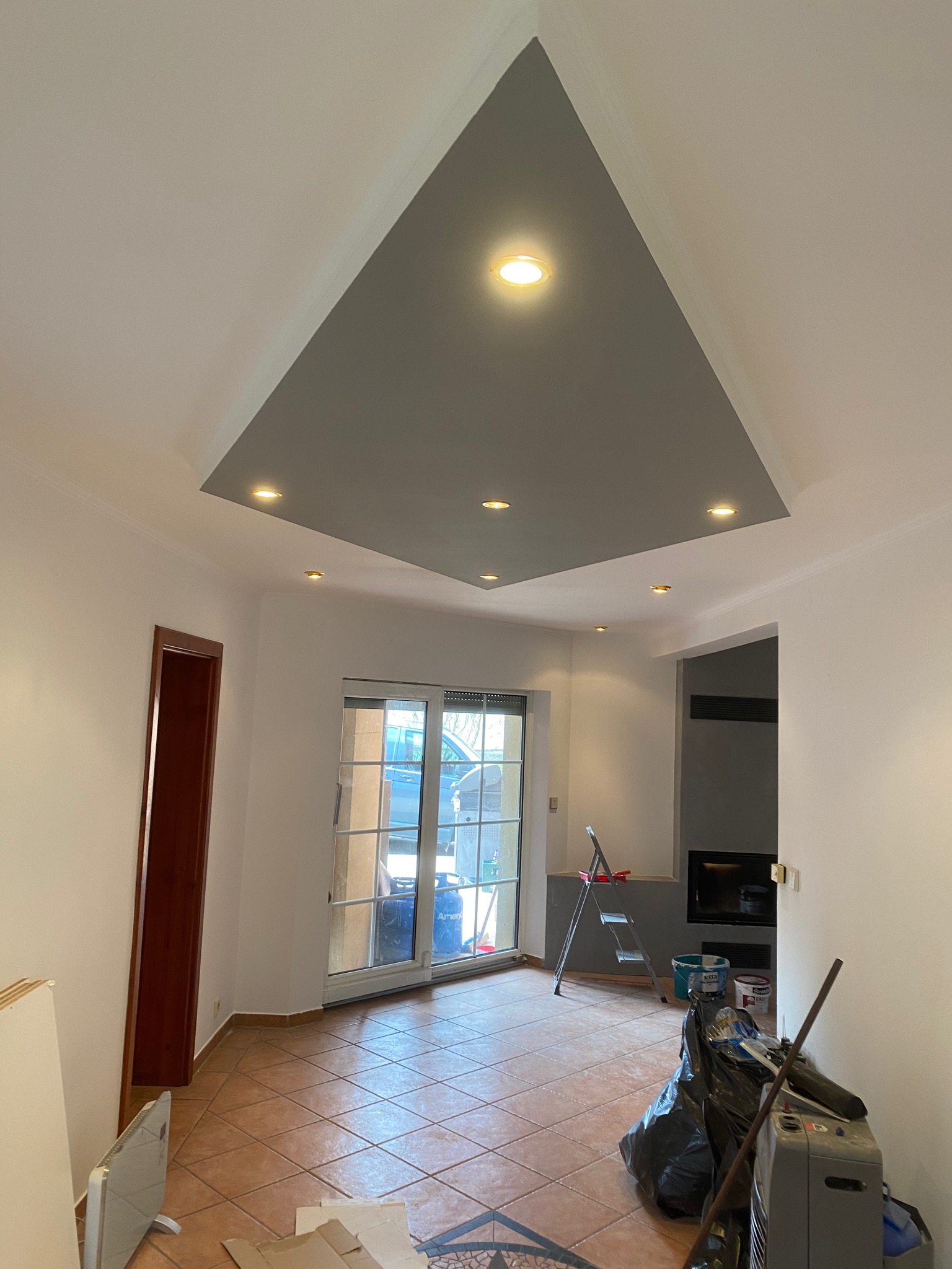 Wnętrze pokoju w trakcie remontu: pomalowany sufit z wbudowanymi punktowymi lampami, drabina, wiadro z farbą i worki na śmieci na podłodze z brązowych płytek.