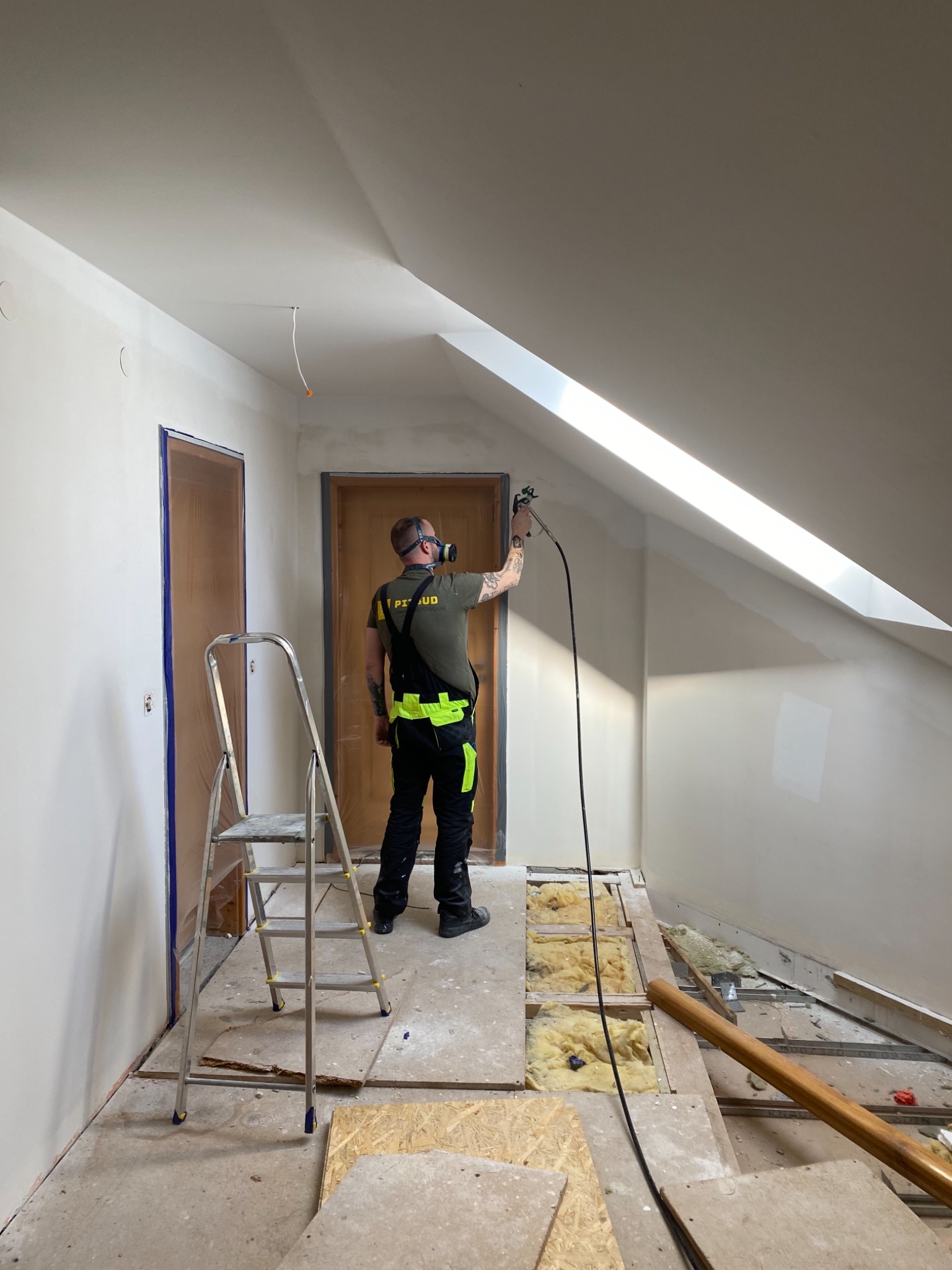 Pracownik w masce ochronnej maluje natryskowo poddasze ze skosem, widoczna drabina i zabezpieczone folią drzwi, podłoga w trakcie remontu.