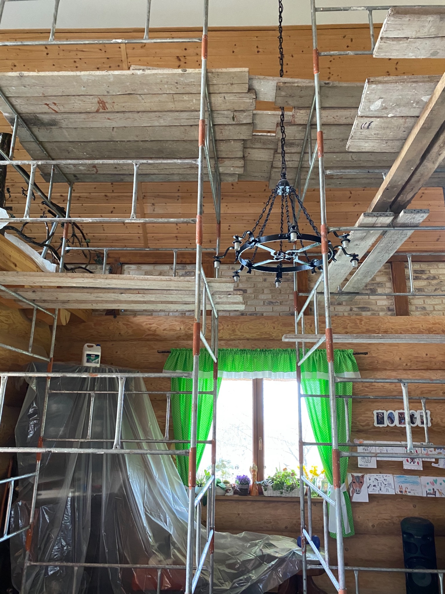 Wnętrze drewnianego domu w trakcie remontu, widoczne rusztowanie, lampa wisząca, okno z zielonymi zasłonami i dziecięce rysunki na ścianie.