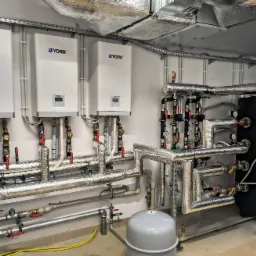 Trzy białe kotły marki York zamontowane na ścianie, z licznymi rurami i zaworami, częściowo izolowanymi termicznie folią aluminiową, oraz czarny zbiornik akumulacyjny w tle.