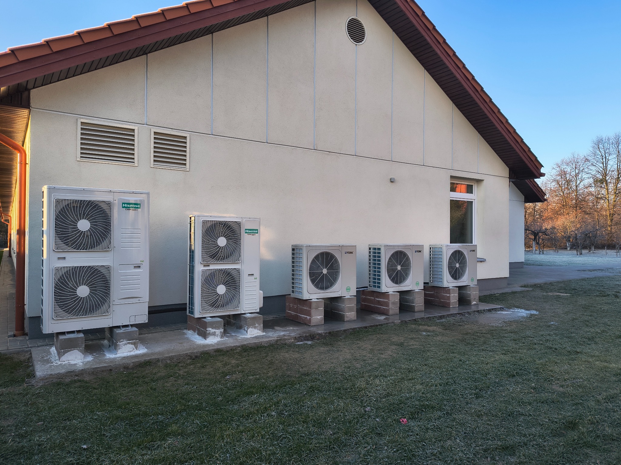 Zewnętrzne jednostki klimatyzacyjne różnych marek (Hisense i York) zamontowane na betonowych podstawach wzdłuż ściany budynku z jasną elewacją i brązowym dachem, trawa pokryta szronem.