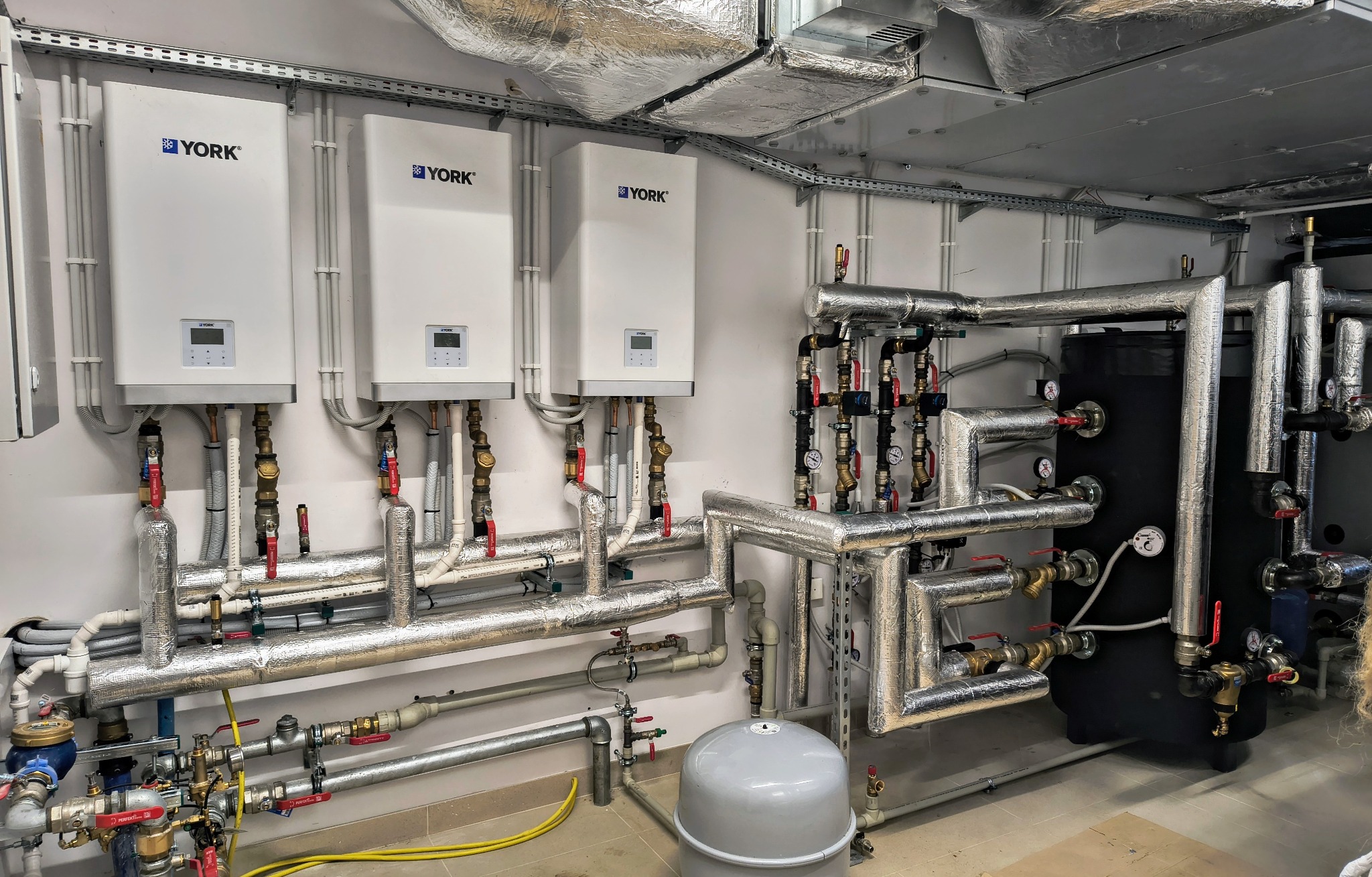Trzy białe kotły marki York zamontowane na ścianie, z licznymi rurami i zaworami, częściowo izolowanymi termicznie folią aluminiową, oraz czarny zbiornik akumulacyjny w tle.