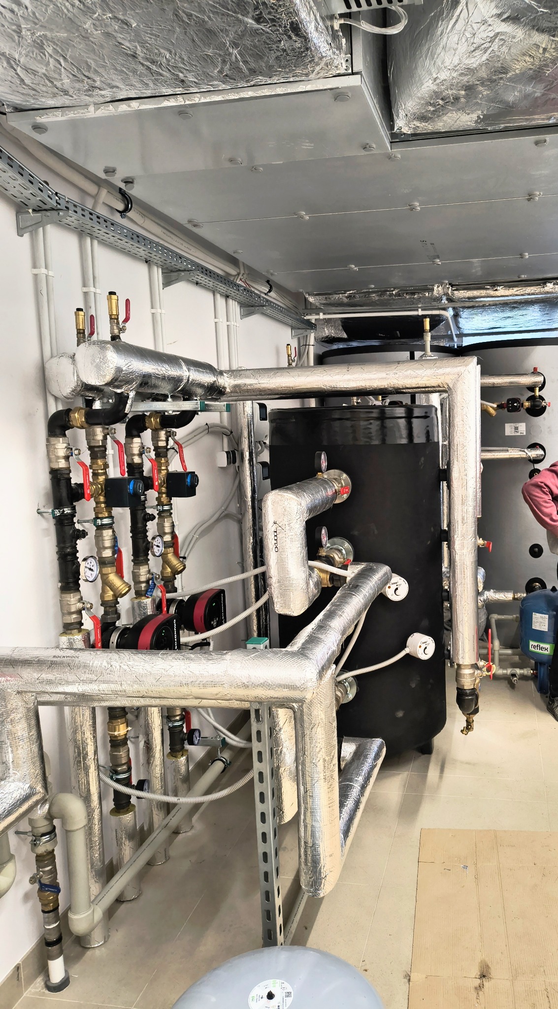 Kotłownia z czarnym zasobnikiem ciepłej wody, rurami w srebrnej izolacji, zaworami i manometrami, widoczne elementy instalacji na suficie.