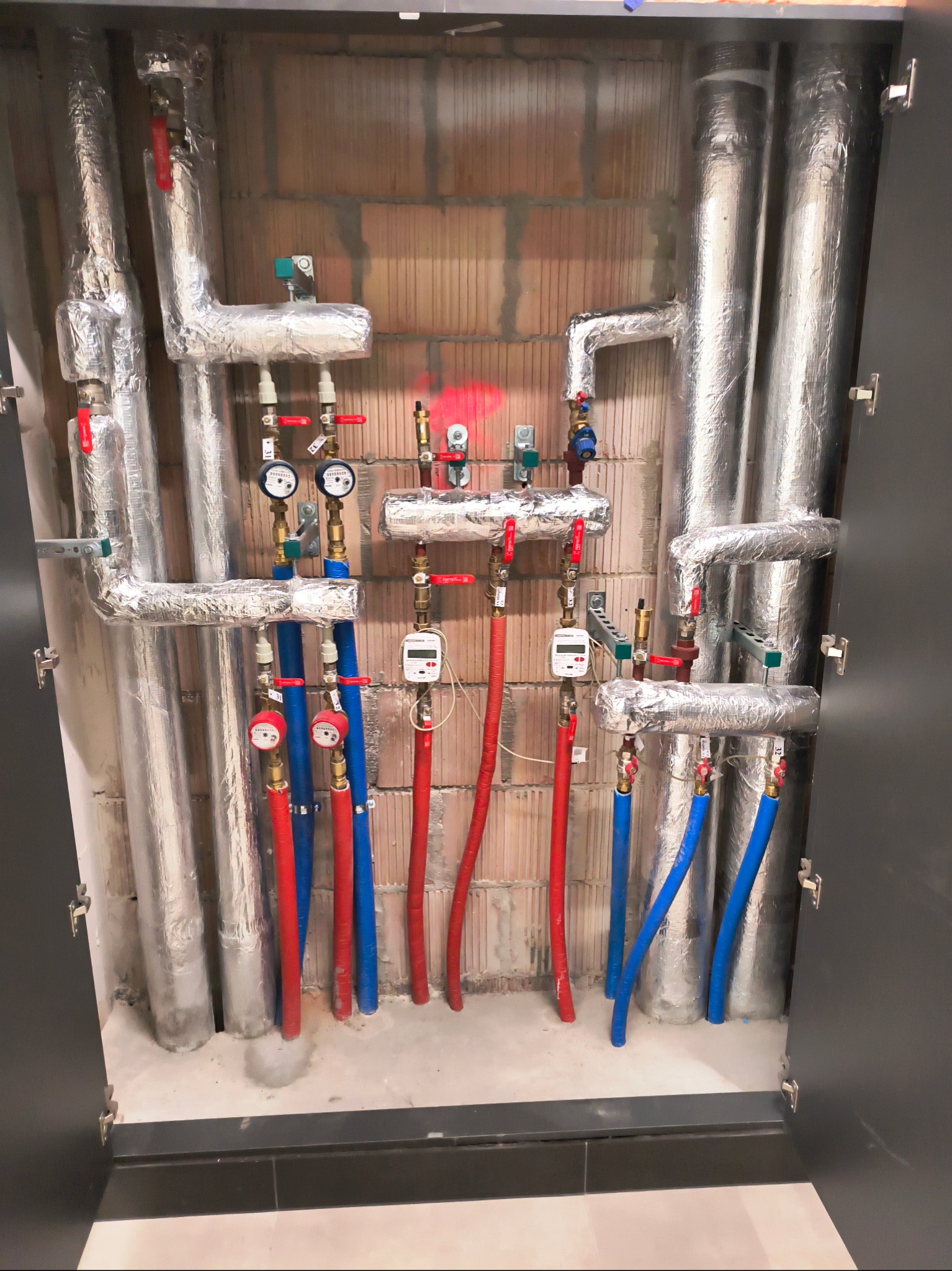 Zabudowana instalacja grzewcza i wodna z rurami w izolacji aluminiowej, zaworami, licznikami ciepła i wodomierzami, na tle ściany z cegły dziurawki.