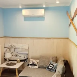 Wnętrze pokoju z klimatyzatorem Samsung na ścianie w stylu marynistycznym, szare sofy z poduszkami w morskie wzory, drewniane wiosła jako dekoracja.