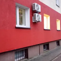 Dwa klimatyzatory zamontowane jeden nad drugim na czerwonej ścianie budynku z widocznymi oknami.