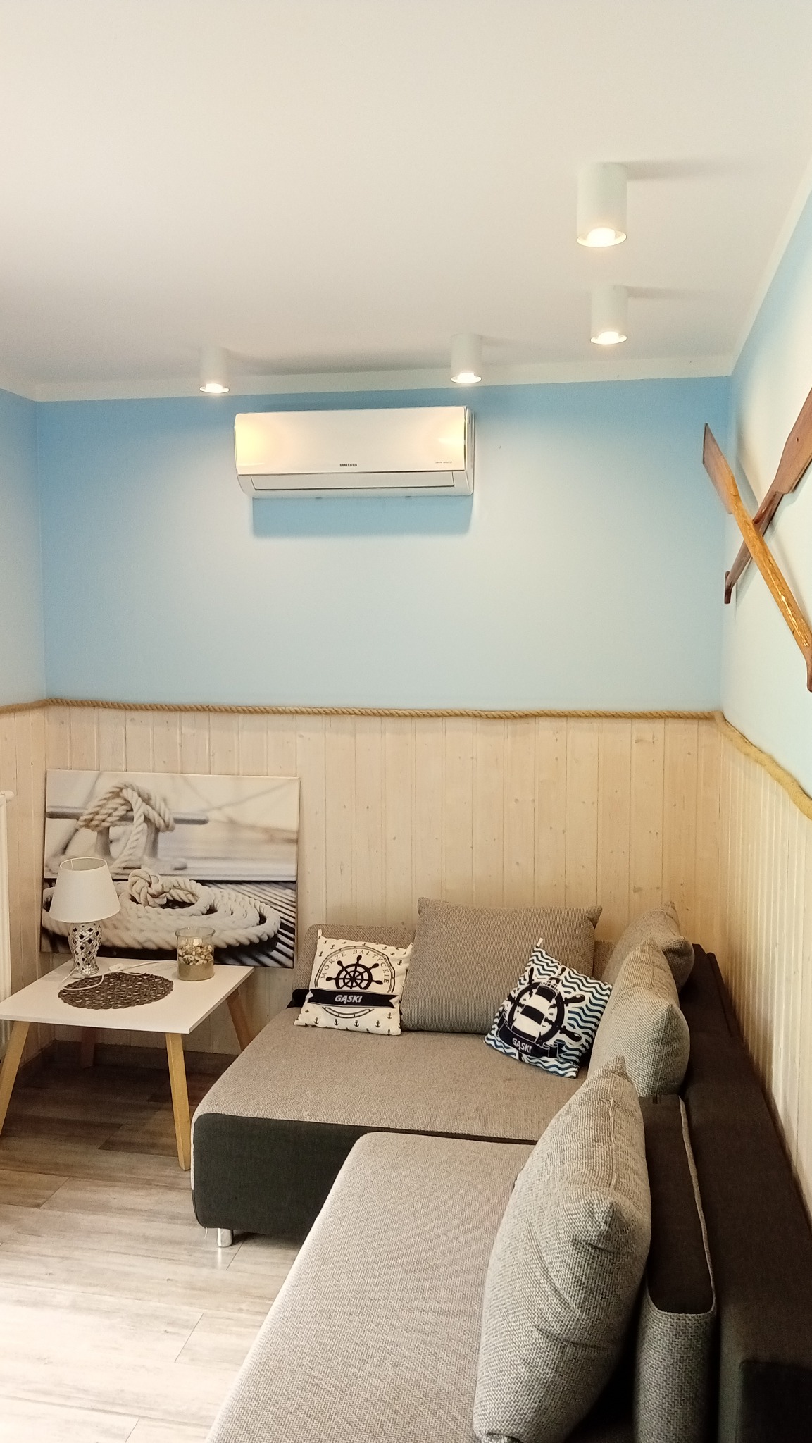 Wnętrze pokoju z klimatyzatorem Samsung na ścianie w stylu marynistycznym, szare sofy z poduszkami w morskie wzory, drewniane wiosła jako dekoracja.