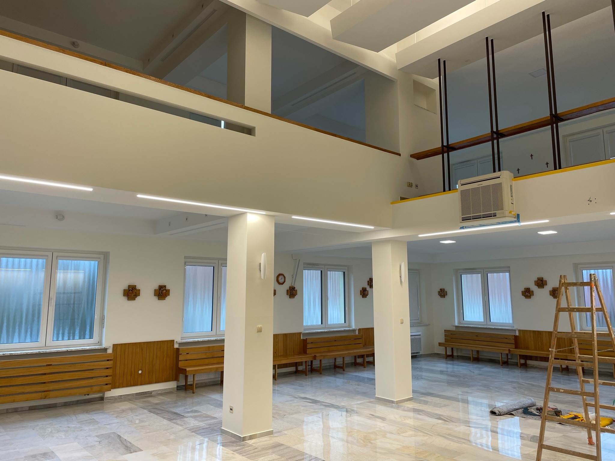 Jasne, przestronne wnętrze po remoncie z marmurową posadzką, widoczne drewniane ławki, drabina malarska i narzędzia, ściany świeżo pomalowane na biało, charakterystyczne krzyże na ścianach.