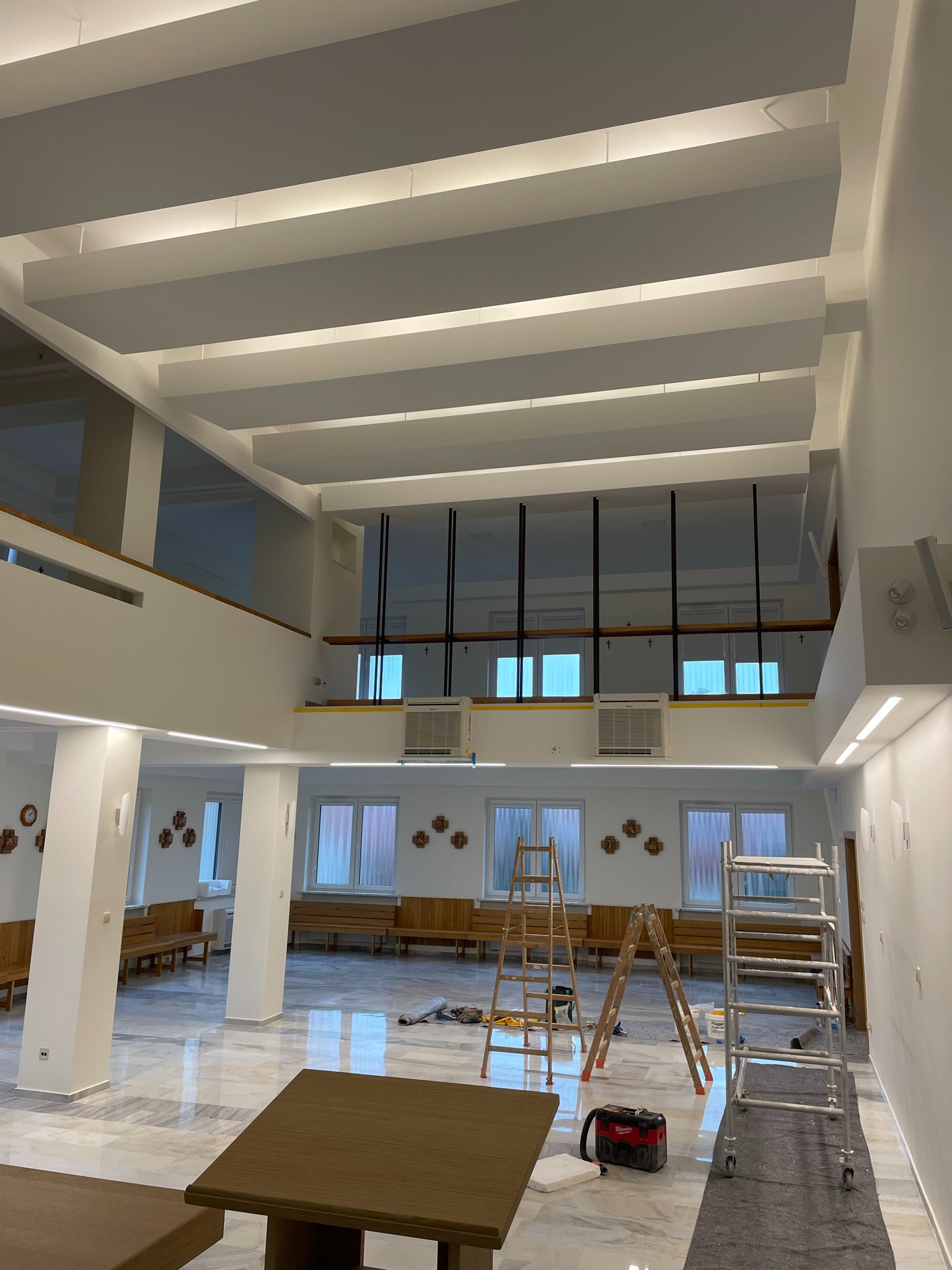 Wysokie wnętrze kościoła w trakcie malowania, widoczne rusztowania, drabiny, marmurowa posadzka i oświetlenie sufitowe w postaci białych belek.