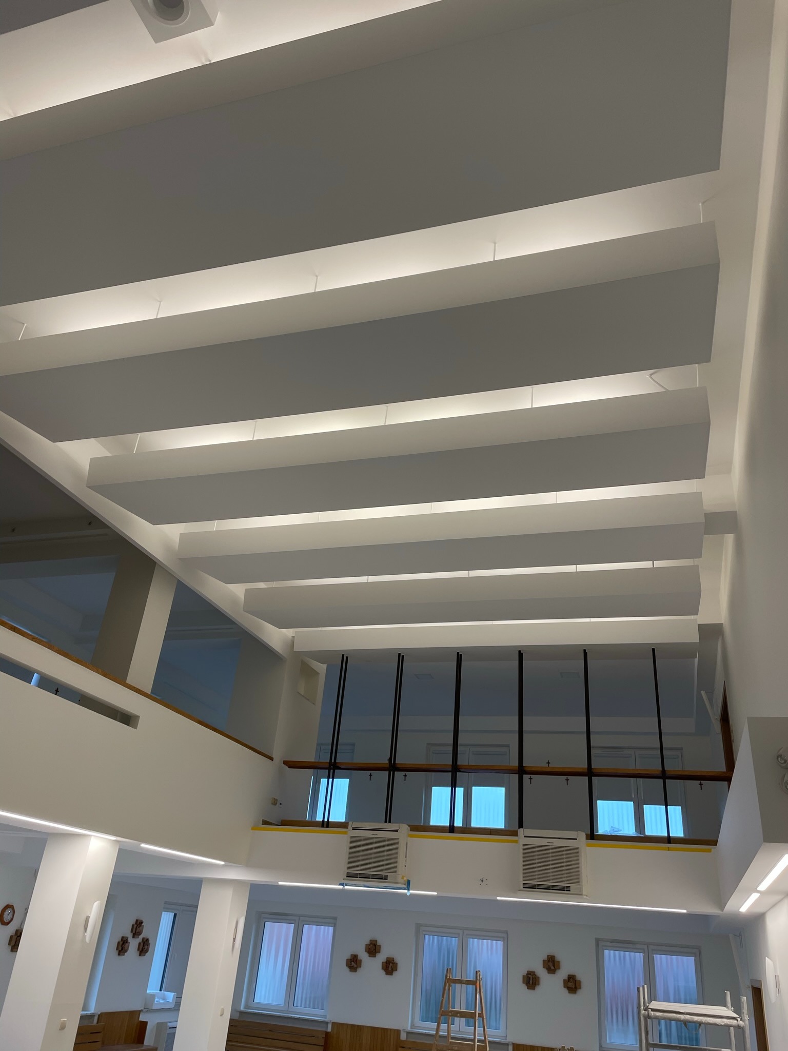 Dwupoziomowe wnętrze z białymi ścianami i sufitem z podwieszanymi panelami oświetleniowymi, widok z dołu na górę, z widoczną drabiną i rusztowaniem na niższym poziomie.