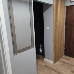 Usługi remontowo wykończeniowe Piotr Sobieraj - Wnętrze przedpokoju po remoncie: lustro w srebrnej ramie na szarej ścianie, drewniana ażurowa ścianka działowa, podłoga z paneli imitujących drewno.