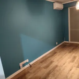 Pomieszczenie po odświeżeniu: turkusowa ściana, drewnopodobna podłoga, biała listwa przypodłogowa, klimatyzator na ścianie, widoczne gniazdka elektryczne.