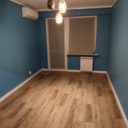 Usługi remontowo wykończeniowe Piotr Sobieraj - Puste pomieszczenie z panelami podłogowymi w odcieniach drewna, pomalowane na niebiesko ściany, biały grzejnik pod oknem i designerska lampa wisząca z trzema kloszami.