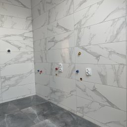Usługi remontowo wykończeniowe Piotr Sobieraj - Wnętrze łazienki w trakcie prac wykończeniowych, widoczne białe płytki imitujące marmur na ścianach oraz szare płytki podłogowe, przygotowane punkty instalacji wodnej z kolorowymi oznaczeniami...