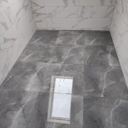 Usługi remontowo wykończeniowe Piotr Sobieraj - Wykończona łazienka z szarymi płytkami imitującymi marmur na podłodze i białymi na ścianach, widoczne odbicie światła w płytkach podłogowych.