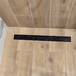 Ukończona łazienka z drewnopodobnymi płytkami, czarnym odpływem liniowym i widocznym fragmentem słuchawki prysznicowej oraz butem pracownika