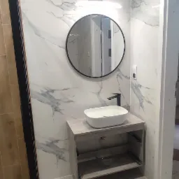 Nowoczesna łazienka z umywalką nablatową na szafce z otwartą półką, okrągłe lustro z podświetleniem LED i marmurowe płytki ścienne.