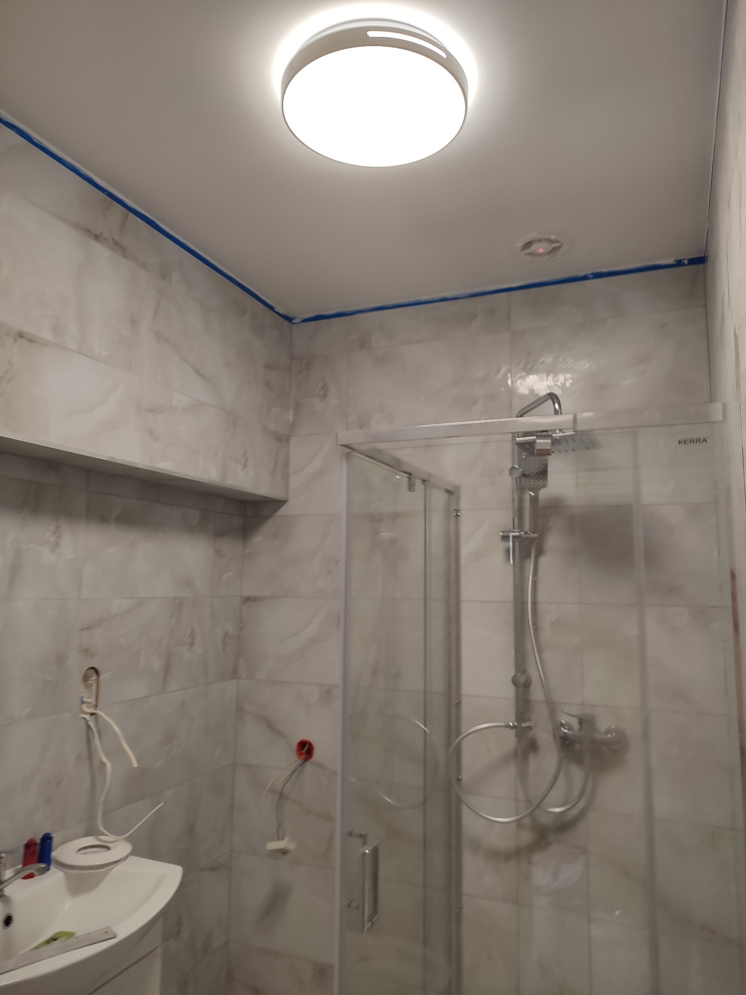 Łazienka w trakcie remontu: widoczne płytki imitujące marmur, kabina prysznicowa ze szklanymi drzwiami, umywalka z baterią, wystające przewody elektryczne ze ściany, sufit z taśmą malarską...