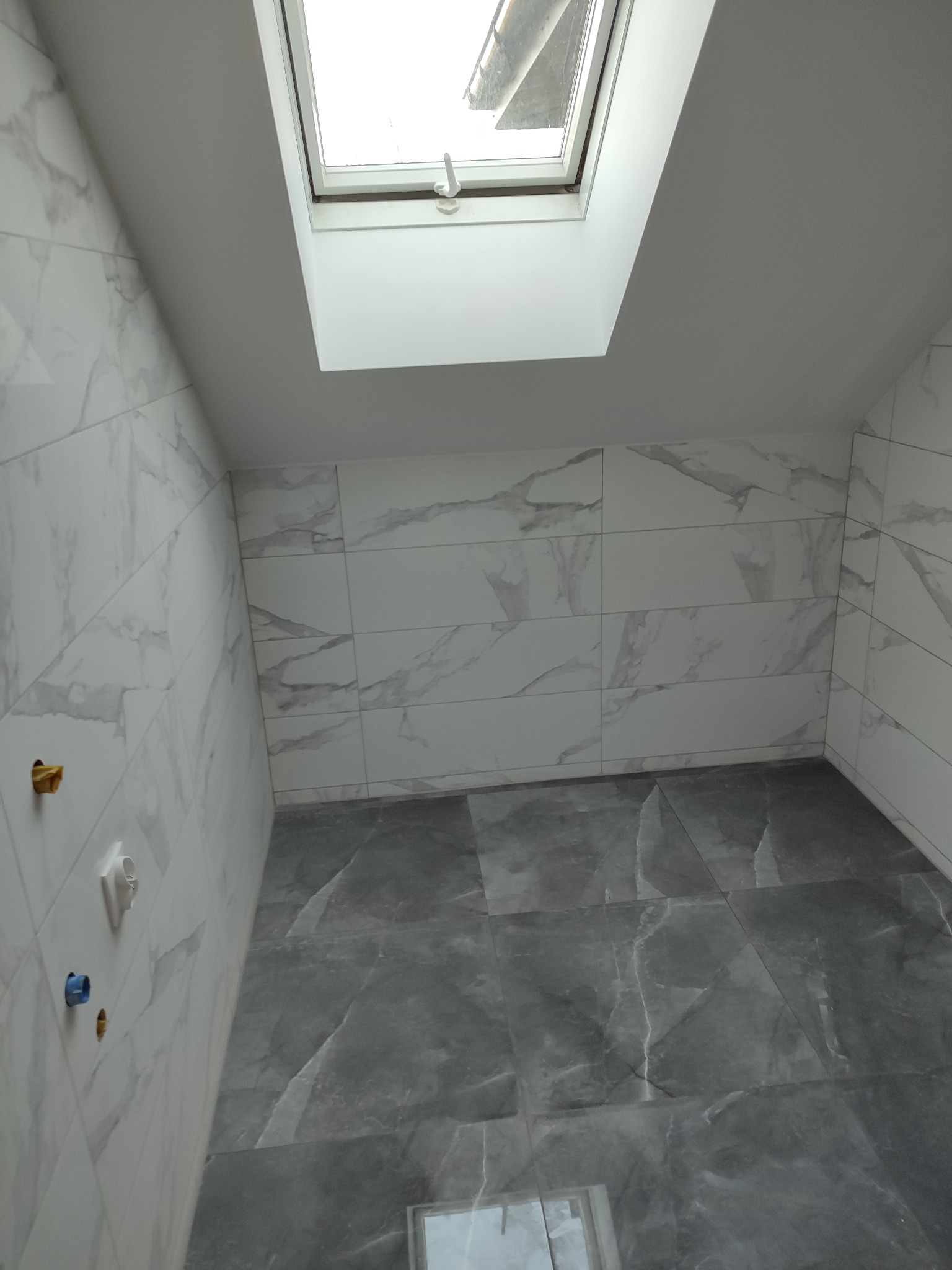 Wykończona łazienka z szarymi płytkami podłogowymi imitującymi marmur i białymi płytkami ściennymi z szarym wzorem marmuru, oknem dachowym i wystającymi przyłączami wodnymi.