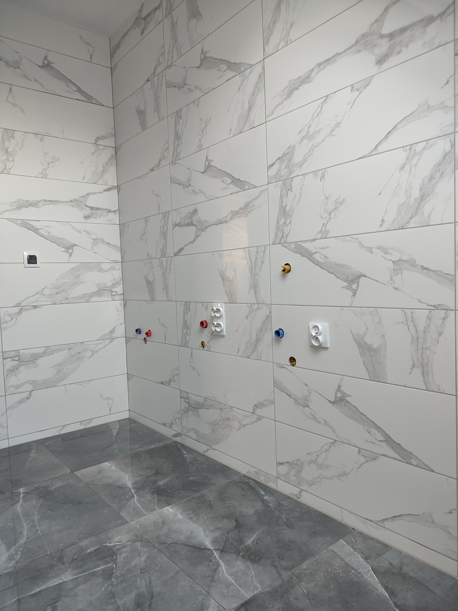 Wnętrze łazienki w trakcie prac wykończeniowych, widoczne białe płytki imitujące marmur na ścianach oraz szare płytki podłogowe, przygotowane punkty instalacji wodnej z kolorowymi oznaczeniami...