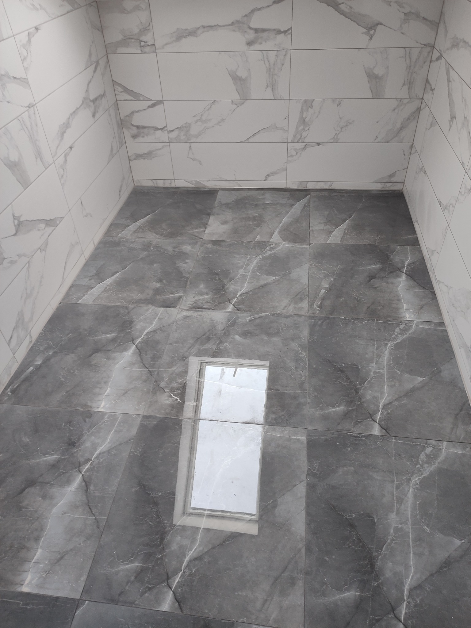 Wykończona łazienka z szarymi płytkami imitującymi marmur na podłodze i białymi na ścianach, widoczne odbicie światła w płytkach podłogowych.