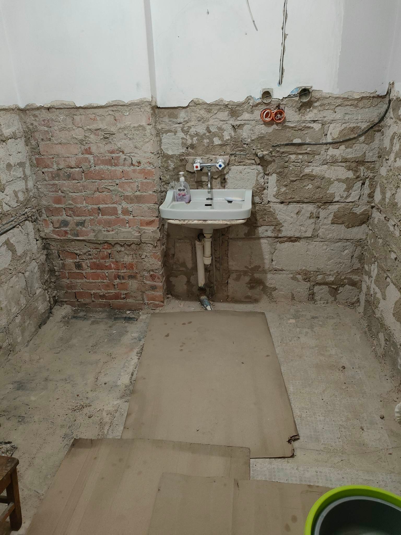 Łazienka w trakcie demontażu, odsłonięte cegły i instalacje, stara umywalka, podłoga zabezpieczona kartonem, widoczne ślady po starych płytkach.