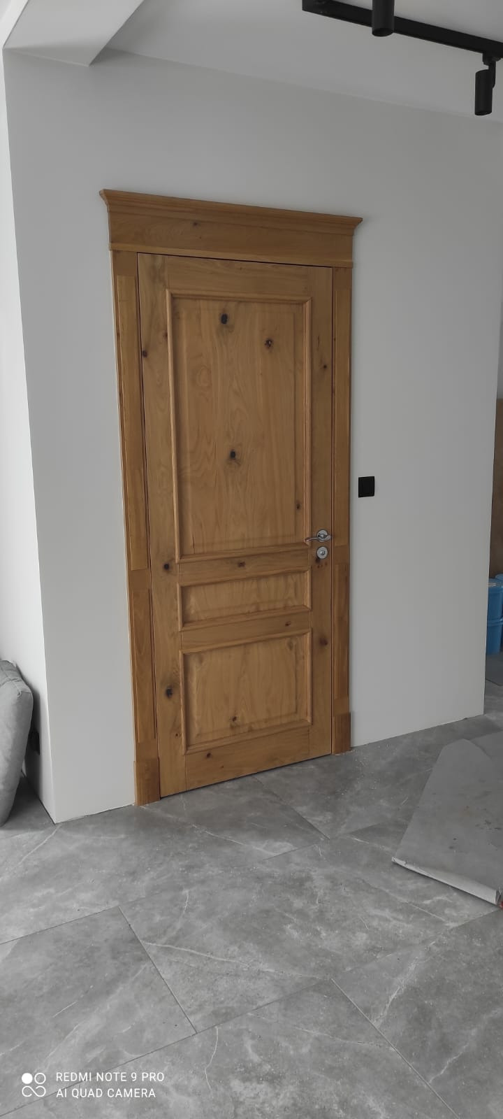 Dębowa trzyskrzydłowa wewnętrzna stolarka drzwiowa z prostokątnymi płycinami, zamontowana w białej ścianie, widoczna na tle szarej podłogi z płytek imitujących marmur.